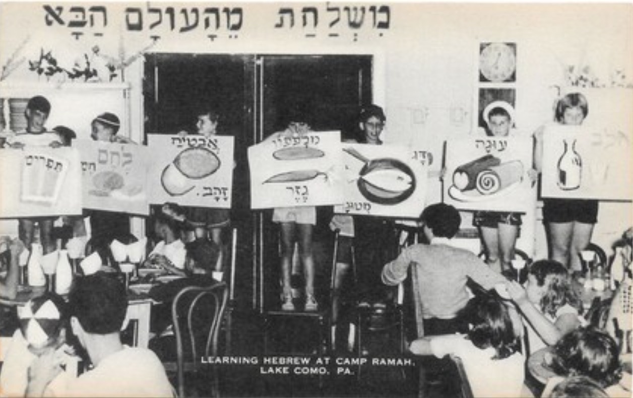 A postcard with a photograph of a group of people holding up signs with Hebrew text and the subtitle "Learning Hebrew at Camp Ramah, Lake Como, PA"